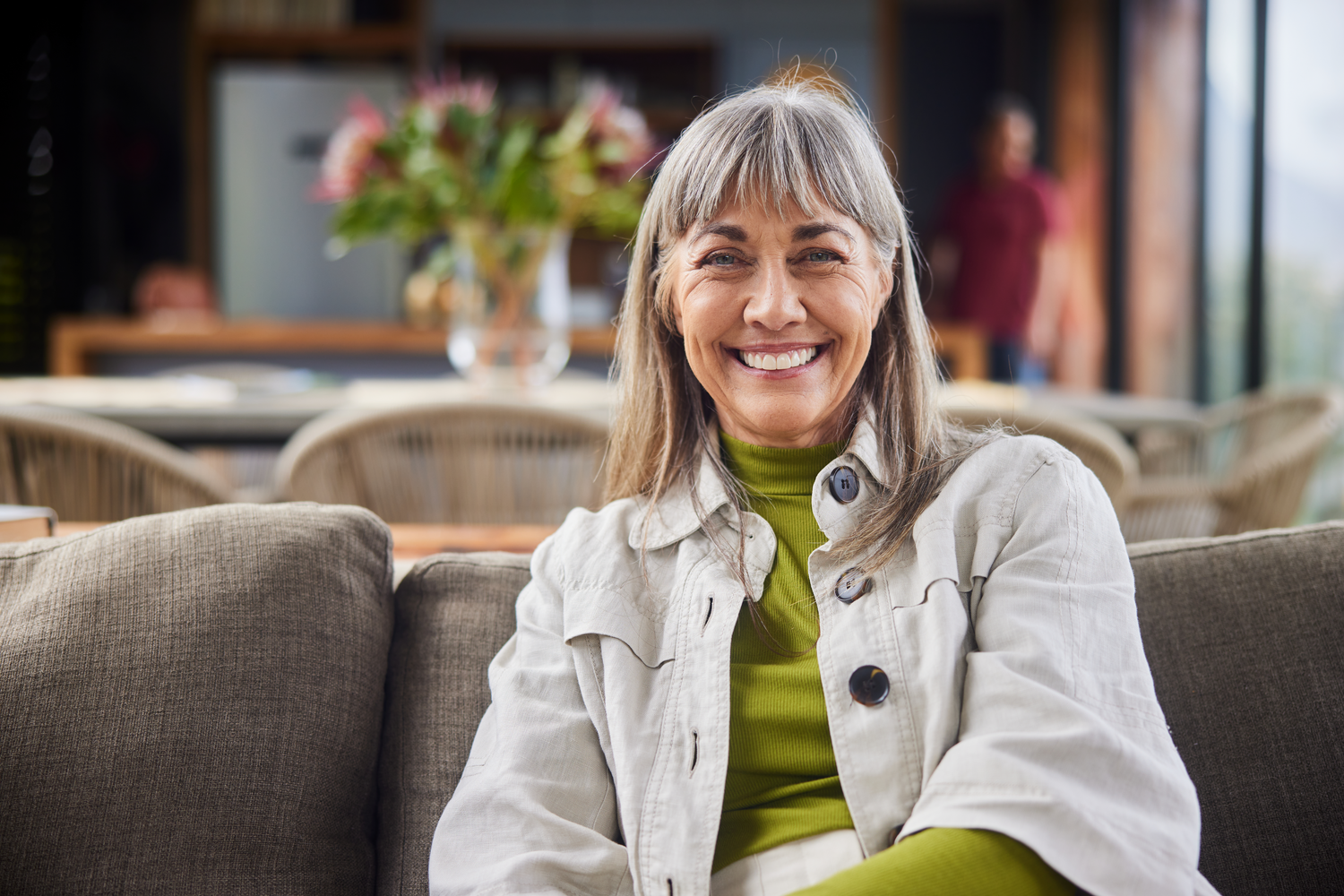 A smiling woman sitting on a couch, wearing a beige jacket and a green top, with a professionally edited life story video interview background.